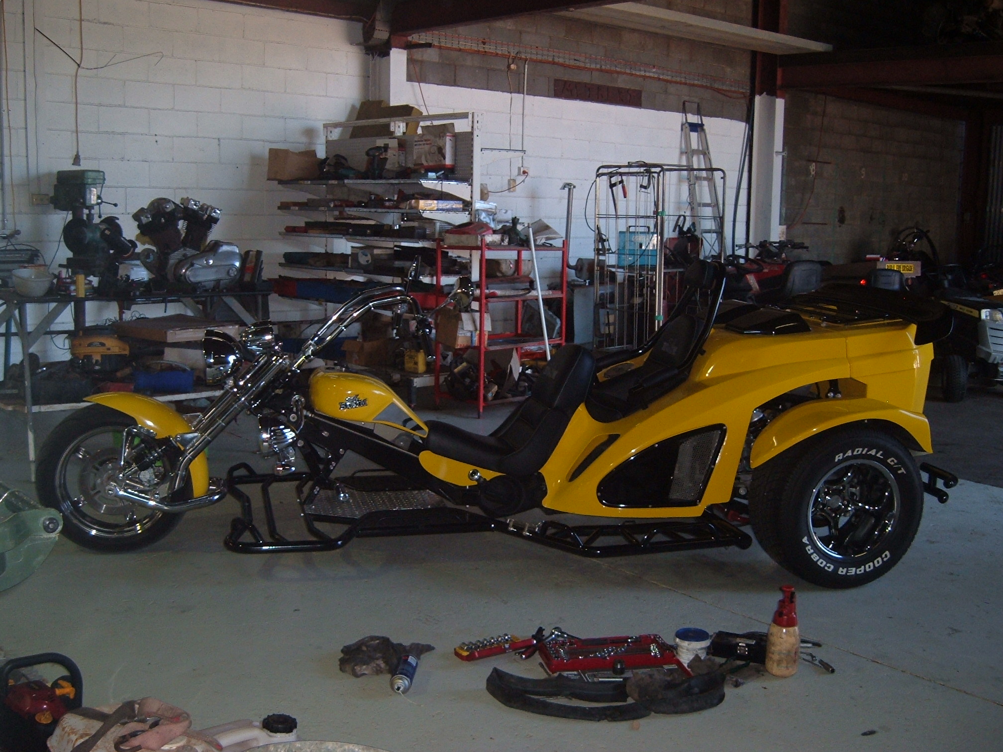 A bright yellow two seat trike parked in the middle of the workshop floor