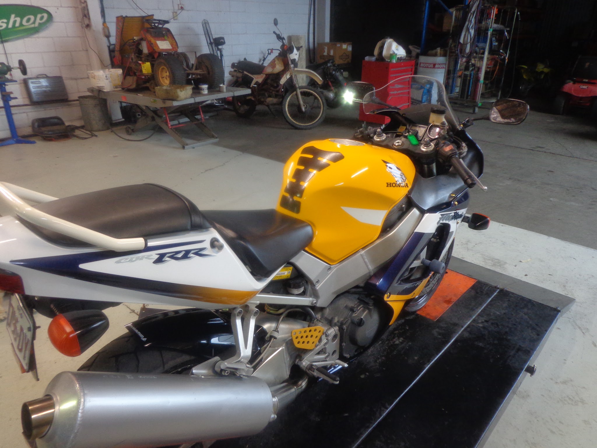 A Honda CBR900RR Fireblade on the workshop ramp, with a quad bike, a small dirt bike and a mower visible behind it