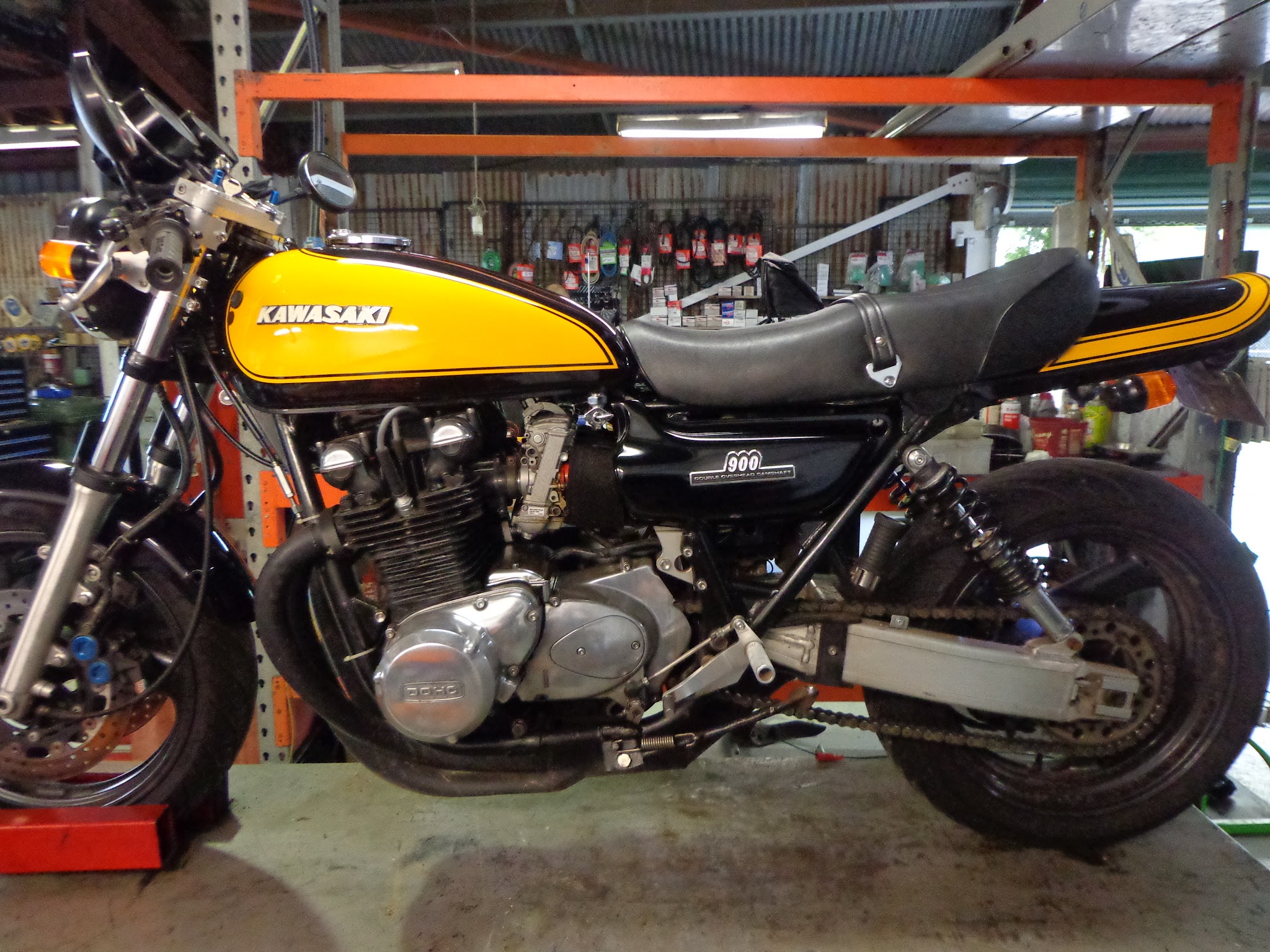 A yellow tanked Kawasaki Z900 photographed side on in front of a rack of drive belts