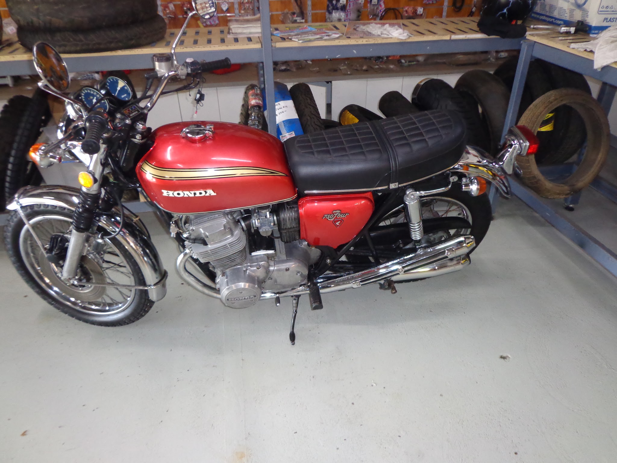 A candy red Honda CB750 Four with gold pinstriping and four chrome exhaust pipes, finished and standing on the workshop floor